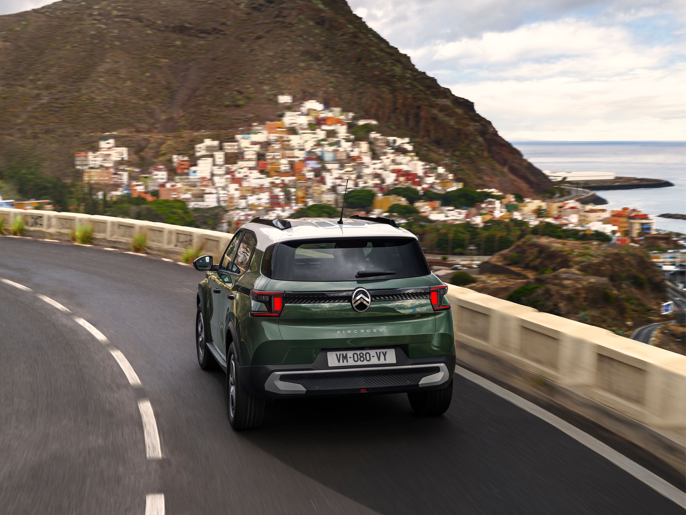 Driekwart achteraanzicht rijdend van de Citroën ë‑C3 Aircross SUV op een kustweg voor zelfverzekerde, soepele en betaalbare dagelijkse mobiliteit.